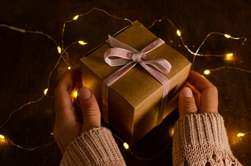 Woman holding wrappd gift box with pink ribbon, garland lights on the wooden surface