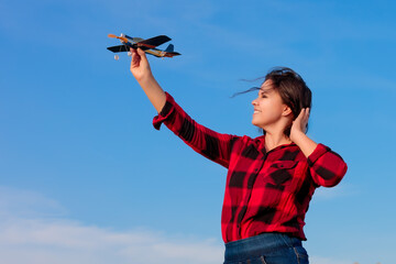A young woman in a red checked shirt and disheveled hairs against the blue sky launches a toy paper airplane by her hand. Image on the theme: tourism and travel, aviation, freedom and inspiration.
