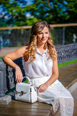 A young girl in a white dress sits in the park on a bench with a white handbag. Summer sunny day