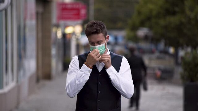 An elegant young man wearing a black waistcoat and white shirt putting on a green protective Covid-19 mask, trying it and then throwing it away contemptuously over his shoulder and smiling, 4k.