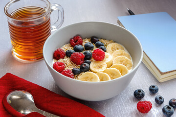 Side view of a healthy breakfast with oatmeal in a bowl, banana slices, raspberry, blueberry, a cup of tea and a notebook with a pen on light grey tabletop
