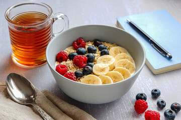 Side view of a healthy breakfast with oatmeal in a bowl, banana slices, raspberry, blueberry, a cup of tea and a notebook with a pen on light grey tabletop