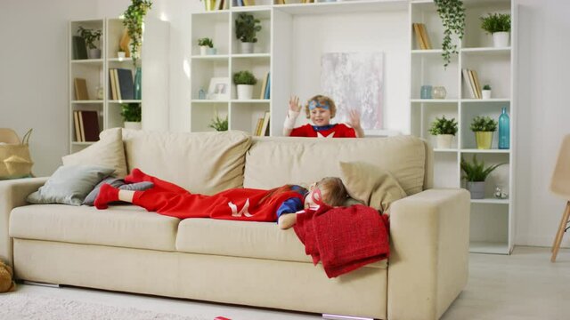 Full shot of small girl laying on sofa in superhero costume pretending sleeping and little boy in blue eye mask scaring her and laughing together