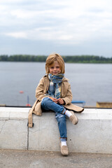 Fototapeta na wymiar blond boy dressed in a coat and scarf sits on a stone border.