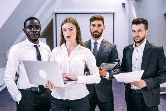 Serious Diverse Professional Business Team Is Standing In The Office, Looking At The Camera, Holding Modern Gadgets And Documents.