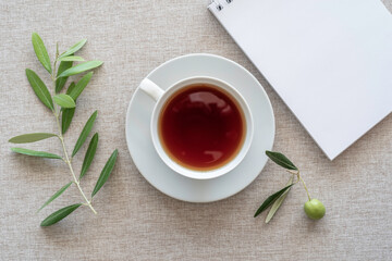 Tea break concept. Tea cup, blank notebook paper, olive branch on cotton fabric background. 