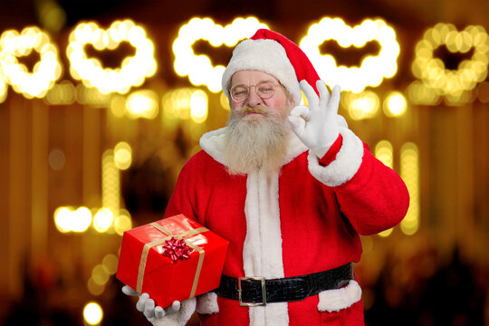 Santa Claus showing okey sign. Cheerful Santa Claus with Christmas present showing ok sign standing on festive shimmering background.