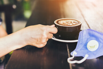 Hot Cappuccino coffee with protective mask on a wooden table for people being quarantined in the situation of the spread of the corona virus (Covid-19).  New normal lifestyle concept. Selective focus