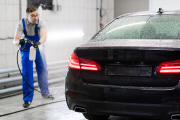 Car with turned on backlights standing in car wash service. Worker cover the car with foam with high-pressure sprayer.