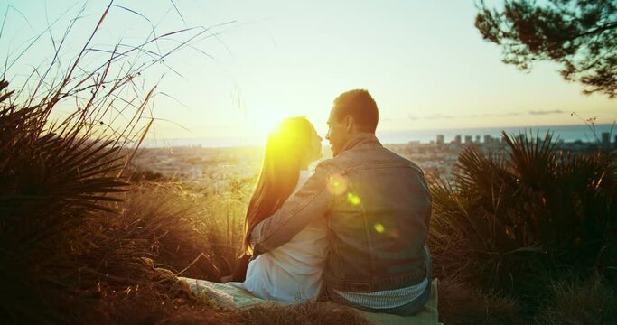 Shot from back of happy young couple kissing on first date sitting at nature hill outdoors, attractive boyfriend embracing pretty girlfriend in love enjoying beautiful sunlight sunset at park