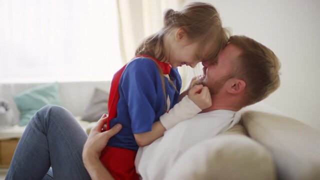 Medium closeup of smiling father embracing little laughing girl in superhero costume tenderly and dancing with pressed foreheads looking at each other softly