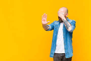 young bald and bearded man covering face with hand and putting other hand up front to stop camera,...