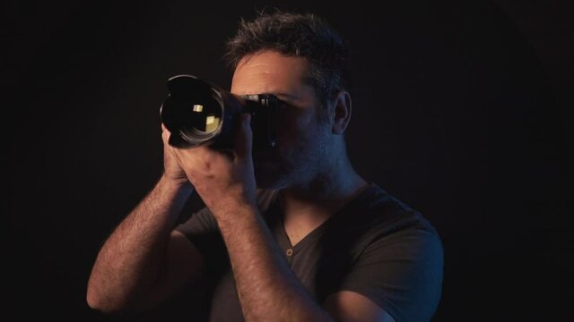 Professional male photographer taking pictures and checking them on the viewfinder to review. Dark studio with black background. Isolated freelance artist working with a DSLR mirrorless camera
