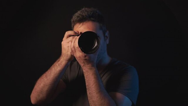 Front view of a Young professional male photographer taking a photo in a dark studio. Black background isolated man shooting a DSLR mirrorless camera. 