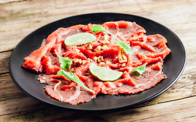 Black plate with carpaccio of beef with baby arugul and lime on rustic wooden background.