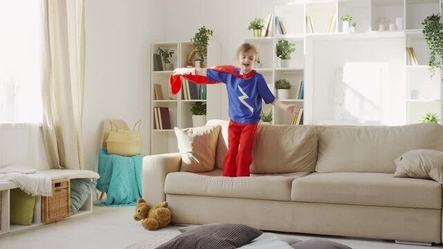 Slowmo follow shot of young girl in superhero costume jumping on sofa and waving cloak finally landing on floor with pillows laughing in living room