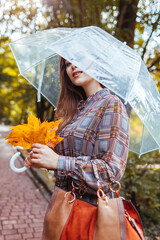 Portrait of young woman walking in park with transparent umbrella holding leaves and handbag. Fall...
