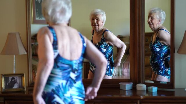 Happy elderly woman looks at herself in mirror as she models a bathing suit she wears in water aerobics class. Concept of active senior female dancing in mirror.