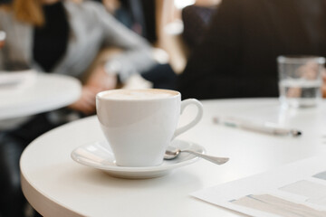 White cup of cappuccino stands on a white table in the interior of a cafe