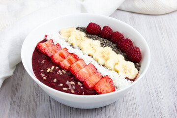 Smoothie bowl or acai berry bowl with chia, strawberries, raspberries, banana slices and coconut flakes. Healthy vegan food. Top view, white wooden background.