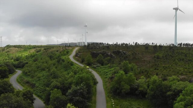 Yellow Car Driving On The Uphill Road Towards The Wind Farm With The Wind Turbines Generating Renewable Energy In Canda, Galicia, Spain. - aerial drone (ascending)