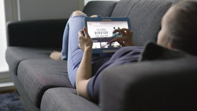 Woman Using Movie Stream Service With Tablet. Choosing Series Or Film And Lying On Couch In Home Living Room. Watching An Episode Of A Tv Show With Mobile Device. Streaming On Demand Video.