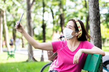 Naklejka premium Woman wearing a face mask sitting using smartphone for take a photo outdoors on a bench in a lush green park. concept of the coronavirus or Covid-19 pandemic.