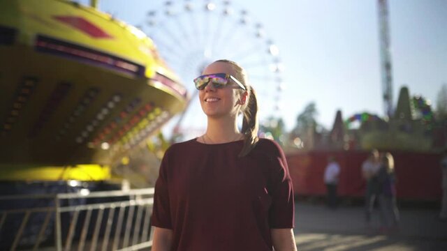 Happy person walking in funfair or amusement park. Young millennial hipster lady enjoying carnival at sunset. Rides, ferris wheel, roller coaster and carousel in the background. Fun summer lifestyle.
