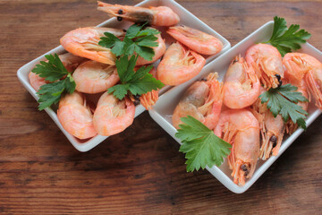 Cooked pink shrimps in the dish served with parsley. Shell-on North Atlantic prawns on the white plates on rustic wooden table. Top view, copy space
