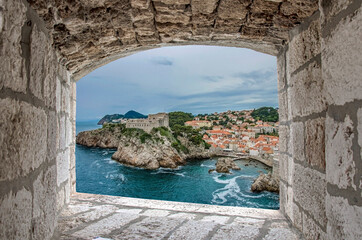 Fototapeta na wymiar View from stone window of Lovrijenac fortress surrounded by blue sea and red rooftops and roofs of historic buildings of Dubrovnik. Beautiful fort on cliffs and rocks.