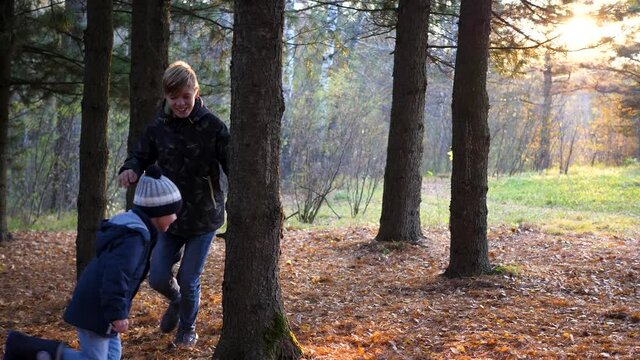 A teenager plays with a child, runs in the Park around a tree and catches up with him. The sun's rays penetrate the pine forest. Laughter and joy of the whole family.