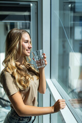Young woman holding a glass of water while looking out of the window at home