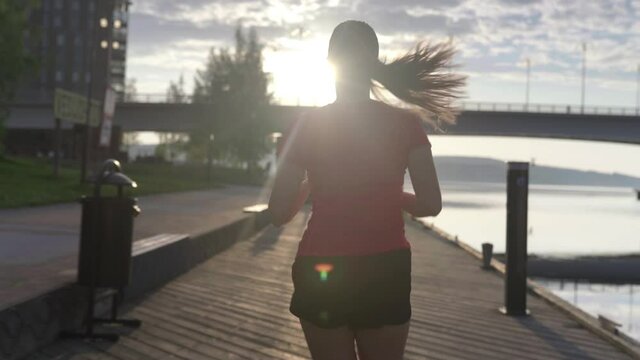 Person running in city. Woman on a morning jog. Following slow motion back view. Fitness runner training. Sunny sky with clouds at sunset or sunrise. Jogger on dock by the lake. Sport motivation.