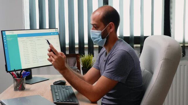 Entrepreneur texing on smartphone wearing mask while working in office room with new normal. Freelancer sitting in modern workspace chatting talking writing using mobile phone with internet technology