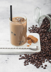 Glass of iced coffee with milk on marble board with jar of coffee beans and salted caramel and long spoon on light background.