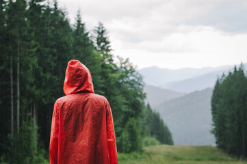 Back view of a male tourist in a bright red raincoat wearing a hood on a rainy day in the mountain fir woods on the hill. Copy space, background. © bodnarphoto