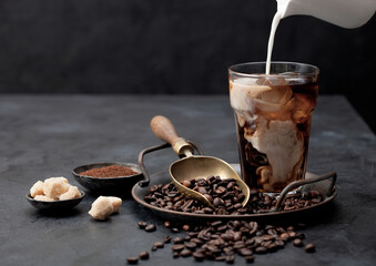 Pouring fresh milk into glass of iced black coffee on tray with beans and ground coffee with cane...