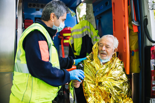 Paramedic rescues an elderly person in distress with an ambulance by covering him with a thermal blanket and making him drink a hot drink - Concept of first aid