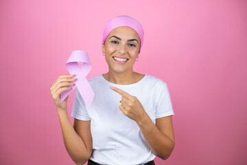 Young beautiful woman wearing pink headscarf holding brest cancer ribbon over isolated pink background smiling happy pointing with hand and finger