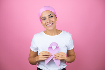 Young beautiful woman wearing pink headscarf holding brest cancer ribbon over isolated pink...