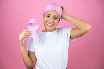 Young beautiful woman wearing pink headscarf holding brest cancer ribbon over isolated pink background confuse and wonder about question. Uncertain with doubt, thinking with hand on head