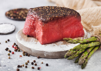 Slice of Raw Beef Topside Joint with Salt and Pepper on round chopping board with asparagus tips and garlic.