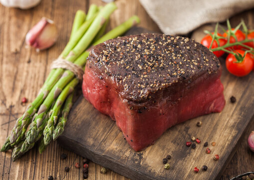 Slice of Raw Beef Topside Joint with Salt and Pepper on wooden chopping board with tomatoes garlic and asparagus tips.