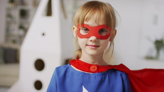 Medium closeup of small girl in superhero costume with red cape waving looking into the distance with cardboard rocket in background