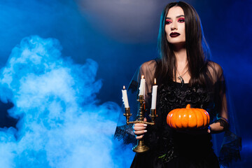 young pale woman with scary makeup holding pumpkin and burning candles on blue with smoke