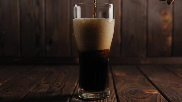 Close-up of dark beer pouring into a glass on a wooden surface, a lot of foam in the beer glass. Slow motion.
