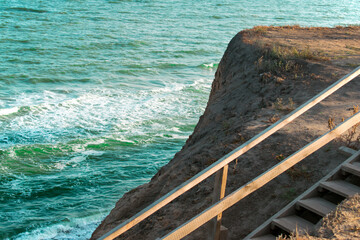 Fototapeta premium Wooden staircase and sea with waves, near a small mountain. Turquoise water color