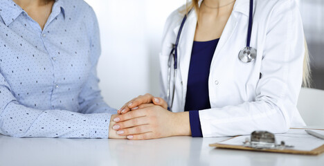 Unknown woman-doctor is holding her patient's hands to reassure a patient, while sitting together at the desk in the cabinet in a clinic. Female physician is using a clipboard and a stethoscope, close