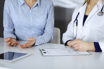 Fototapeta premium Unknown woman- doctor is listening to her patient, while sitting together at the desk in the cabinet in a clinic. Female physician with a stethoscope is writing at clipboard, close up. Perfect medical