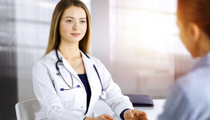 Young beautiful woman-doctor and her patient are discussing patient's current health examination, while sitting together at the desk in the sunny cabinet in a clinic. Female physician is writing some